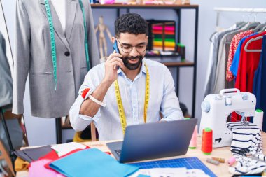 Young arab man tailor talking on smartphone using laptop at tailor shop