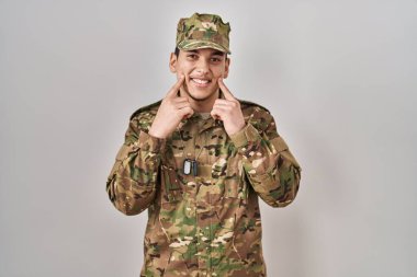 Young arab man wearing camouflage army uniform smiling with open mouth, fingers pointing and forcing cheerful smile 