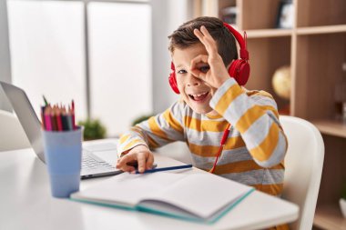 Genç beyaz çocuk evde ödev yapıyor gülümsüyor mutlu bir şekilde tamam işareti yapıyor parmaklarının arasından bakıyor. 