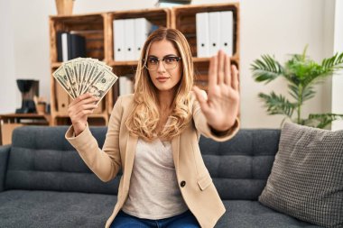 Terapi ofisinde çalışan genç, sarışın bir terapist. Elinde para olan, ciddi ve kendinden emin bir ifadeyle dur işareti yapan, savunma jesti. 