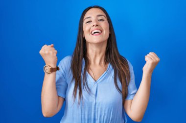 Mavi arka planda dikilen genç esmer kadın kolları havada ve gözleri kapalı bir şekilde başarıyı kutluyor ve şaşırıyor. Kazanan konsept. 