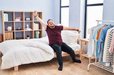 Young hispanic man waking up sitting on bed at bedroom