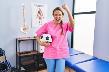Fizyoterapi kliniğinde çalışan genç İspanyol bir kadın futbol topunu tutarken sinirlenmiş ve sinirlenmiş bir şekilde bağırıyor. Öfkeyle bağırıyor ve el kaldırıyor. 