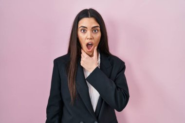 Pembe arka planda iş tarzı giyinen genç esmer kadın elleri çenesinde olan inançsızlıktan, şaşkınlıktan ve şaşkınlıktan büyülenmiş görünüyor. 