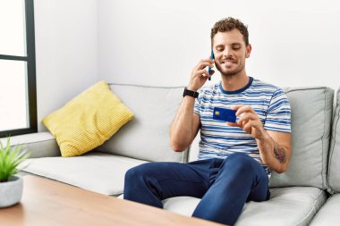 Genç İspanyol adam elinde kredi kartıyla akıllı telefondan konuşuyor.