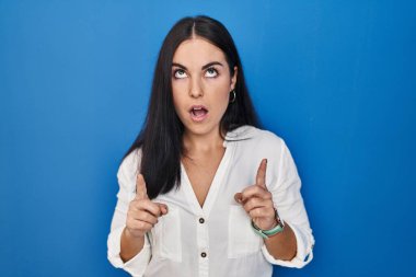 Mavi arka planda duran genç İspanyol kadın şaşırmış ve şaşırmış bir şekilde yukarı bakıyor ve parmaklarıyla işaret ediyor ve kollarını kaldırıyor.. 