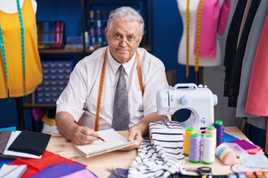 Orta yaşlı, gri saçlı, sırıtan, kendine güvenen, terzi dükkanındaki not defterine çizim yapan bir adam.