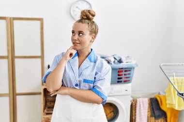 Young caucasian woman wearing cleaner uniform at the laundry room with hand on chin thinking about question, pensive expression. smiling with thoughtful face. doubt concept. 