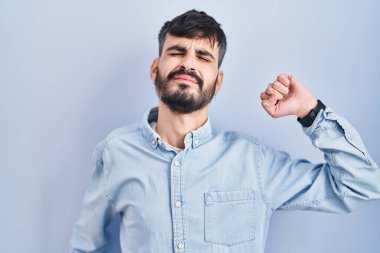 Mavi arka planda dikilen sakallı genç, yorgun ve rahat, uykulu ve sabahın erken saatleri için esniyor. 