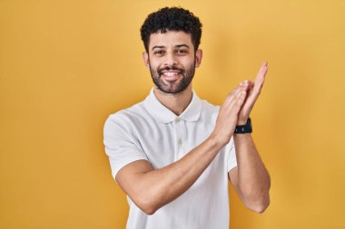 Arap adam sarı arka planda ayakta alkışlıyor ve mutlu ve neşeli, gururlu elleri alkışlıyor. 