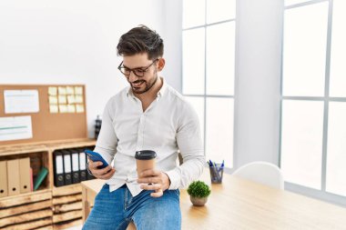 Young hispanic businessman using smartphone and drinking coffee at the office.