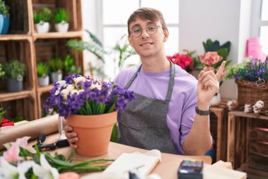 Caucasian blond man working at florist shop with a big smile on face, pointing with hand finger to the side looking at the camera. 