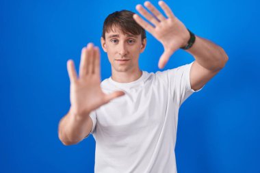 Caucasian blond man standing over blue background doing frame using hands palms and fingers, camera perspective 