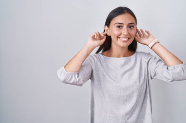 Young hispanic woman standing over white background smiling pulling ears with fingers, funny gesture. audition problem 