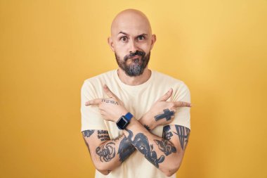 Sarı arka planda dikilen İspanyol dövmeli adam her iki tarafı da parmaklarıyla işaret ediyor, farklı yönlere. 