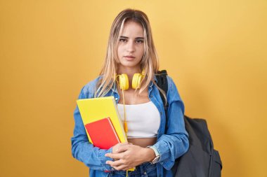 Young blonde woman wearing student backpack and holding books relaxed with serious expression on face. simple and natural looking at the camera. 