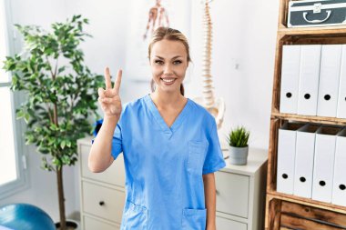 Ağrı kesici kliniğinde çalışan beyaz bir kadın iki numarayı gösterip işaret ederken kendinden emin ve mutlu bir şekilde gülümsüyor.. 