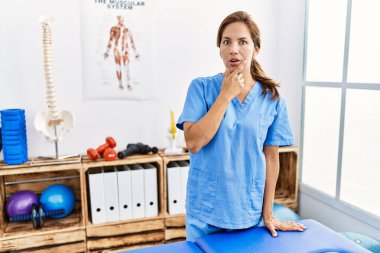 Middle age hispanic physiotherapist woman working at pain recovery clinic looking fascinated with disbelief, surprise and amazed expression with hands on chin 