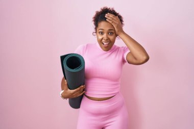 Young hispanic woman with curly hair holding yoga mat over pink background crazy and scared with hands on head, afraid and surprised of shock with open mouth 