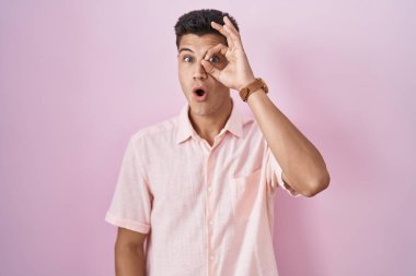 Pembe arka planda duran genç İspanyol adam şaşırmış yüz ifadesiyle şaşırmış, parmaklarının arasından bakan bir jest yapıyor. İnançsız ifade. 