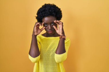 African young woman standing over yellow studio trying to open eyes with fingers, sleepy and tired for morning fatigue 