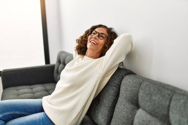 Orta yaşlı İspanyol kadın evde gözlük takarak gülümsüyor.