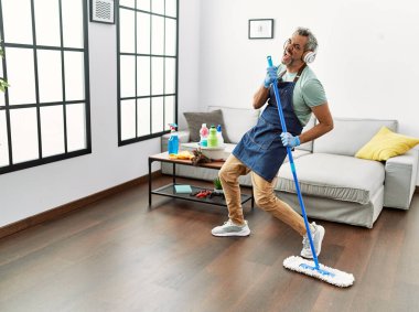 Orta yaşlı, kır saçlı bir adam evde mikrofon olarak paspas kullanarak şarkı söylüyor.