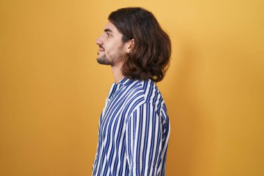 Hispanic man with long hair standing over yellow background looking to side, relax profile pose with natural face with confident smile. 