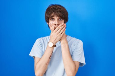 Mavi arka planda duran Latin kökenli genç adam yanlışlıkla elleri ile ağzını kapatırken şok oldu. gizli kavram. 