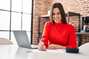 Young beautiful hispanic woman using laptop writing on document at home