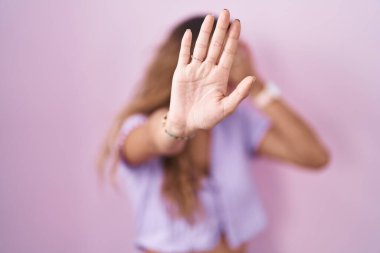 Pembe arka planda duran genç İspanyol kadın elleriyle gözlerini kapatıyor ve üzgün ve korku ifadesiyle dur hareketi yapıyor. utangaç ve olumsuz kavram. 