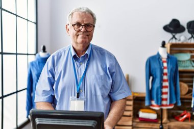 Orta yaşlı, gri saçlı, gülümseyen, kendine güvenen bir adam giyim mağazasında çalışıyor.