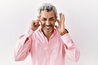 Orta yaşlı İspanyol bir adam izole edilmiş arka planda duruyor. Yüksek sesli müziğin gürültüsü için parmaklarıyla kulaklarını kapatıyor. sağır kavramı. 