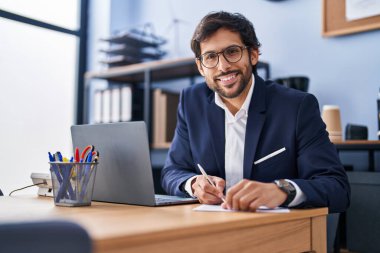 Young hispanic man business worker using laptop write on document at office