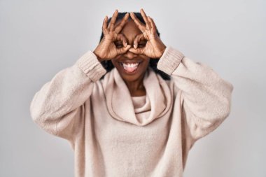 Beyaz arka planda dikilen Afrikalı bir kadın dürbün gibi jest yapıyor. Dürbün dilini çıkarıyor, gözler parmaklarından bakıyor. çılgın ifade. 