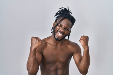 Rastalı saçlı Afrikalı adam izole edilmiş arka planda üstsüz duruyor çok mutlu ve heyecanlı bir şekilde kollarını kaldırıp, gülümseyerek ve başarı için çığlık atarak kazanan bir jest yapıyor. kutlama konsepti. 