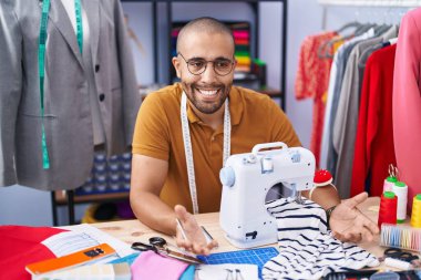 Young latin man tailor sitting on table speaking at atelier