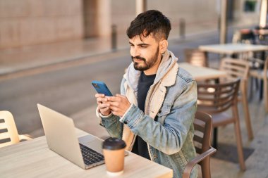 Laptop ve akıllı telefon kullanan İspanyol bir adam kahve dükkanının terasında oturuyor.