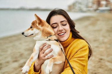 Güzel genç bir kadın sahilde mutlu Shiba Inu köpeğine sarılıyor.