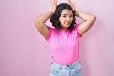Young hispanic woman standing over pink background doing bunny ears gesture with hands palms looking cynical and skeptical. easter rabbit concept. 
