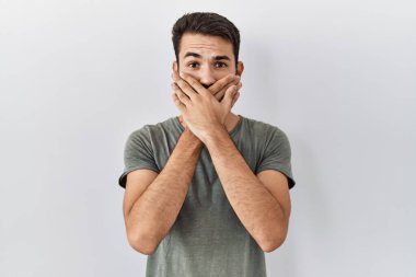Young hispanic man with beard wearing casual t shirt over white background shocked covering mouth with hands for mistake. secret concept. 