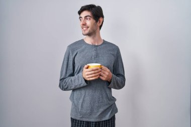 Young hispanic man wearing pajama drinking a cup of coffee looking away to side with smile on face, natural expression. laughing confident. 