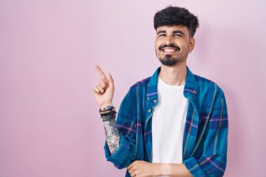 Pembe arka planda duran sakallı, yüzünde kocaman bir gülümseme olan, eli ve parmağı kameraya bakan genç İspanyol bir adam.. 