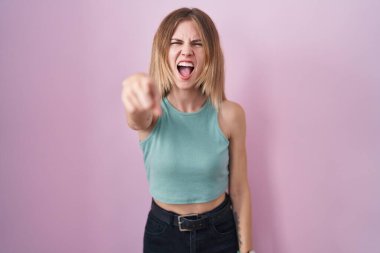 Blonde caucasian woman standing over pink background pointing displeased and frustrated to the camera, angry and furious with you 