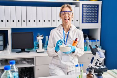 Young caucasian woman working at scientist laboratory sticking tongue out happy with funny expression. 