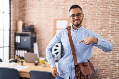Young hispanic man working at the office holding bike helmet smiling happy pointing with hand and finger 