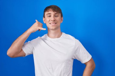 Caucasian blond man standing over blue background smiling doing phone gesture with hand and fingers like talking on the telephone. communicating concepts. 