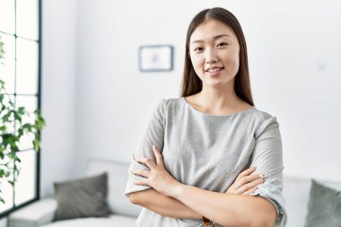 Genç Asyalı kadın oturma odasında kollarını kavuşturup gülümsüyor.