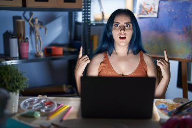 Young modern girl with blue hair sitting at art studio with laptop at night amazed and surprised looking up and pointing with fingers and raised arms. 