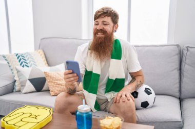 Genç kızıl saçlı adam evde akıllı telefon kullanarak futbol takımını destekliyor.